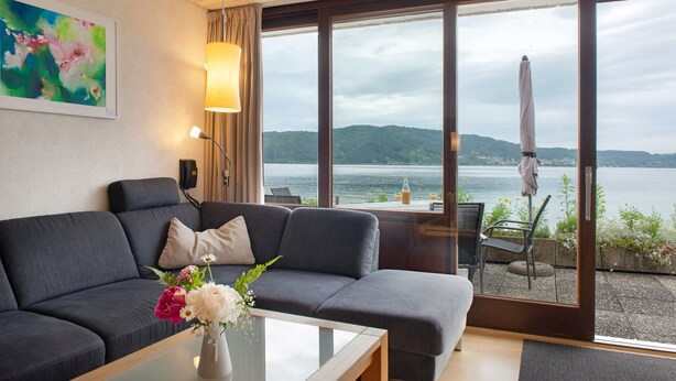A modern living room with a sofa, a coffee table with flowers, a lamp, and a large window with a view of a lake and hills. - Apartment '1, 70sqm' with Large Terrace Right on the Lake (Bodman-Ludwigshafen)