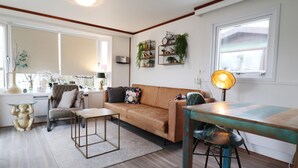 Een woonkamer met een bank, een salontafel, een eettafel en een stoel. Er zijn ramen met jaloezieën, een lamp en een plank met planten.