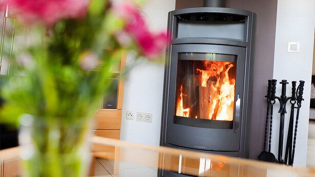 Ein moderner Kaminofen in einem Raum mit freiem Blick auf das Feuer, eine Blumenvase und ein Satz Metallwerkzeuge an der Wand.
