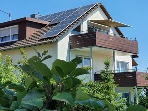 Una casa moderna de dos pisos con paneles solares, un balcón y abundante vegetación al frente.