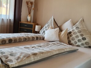 Una cama doble con colcha estampada, una mesita de noche con lámpara, y un florero con flores secas en la pared.