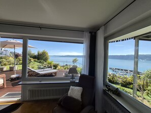 A room with a view of a lake, a chair, a table, and a window with a curtain.