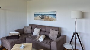 A living room with a brown sofa, a painting of a beach scene, a floor lamp, and a small table with a tissue box. - Apartment 'Panorama-seetraum' with Lake View, Private Terrace and Wi-Fi (Sipplingen)
