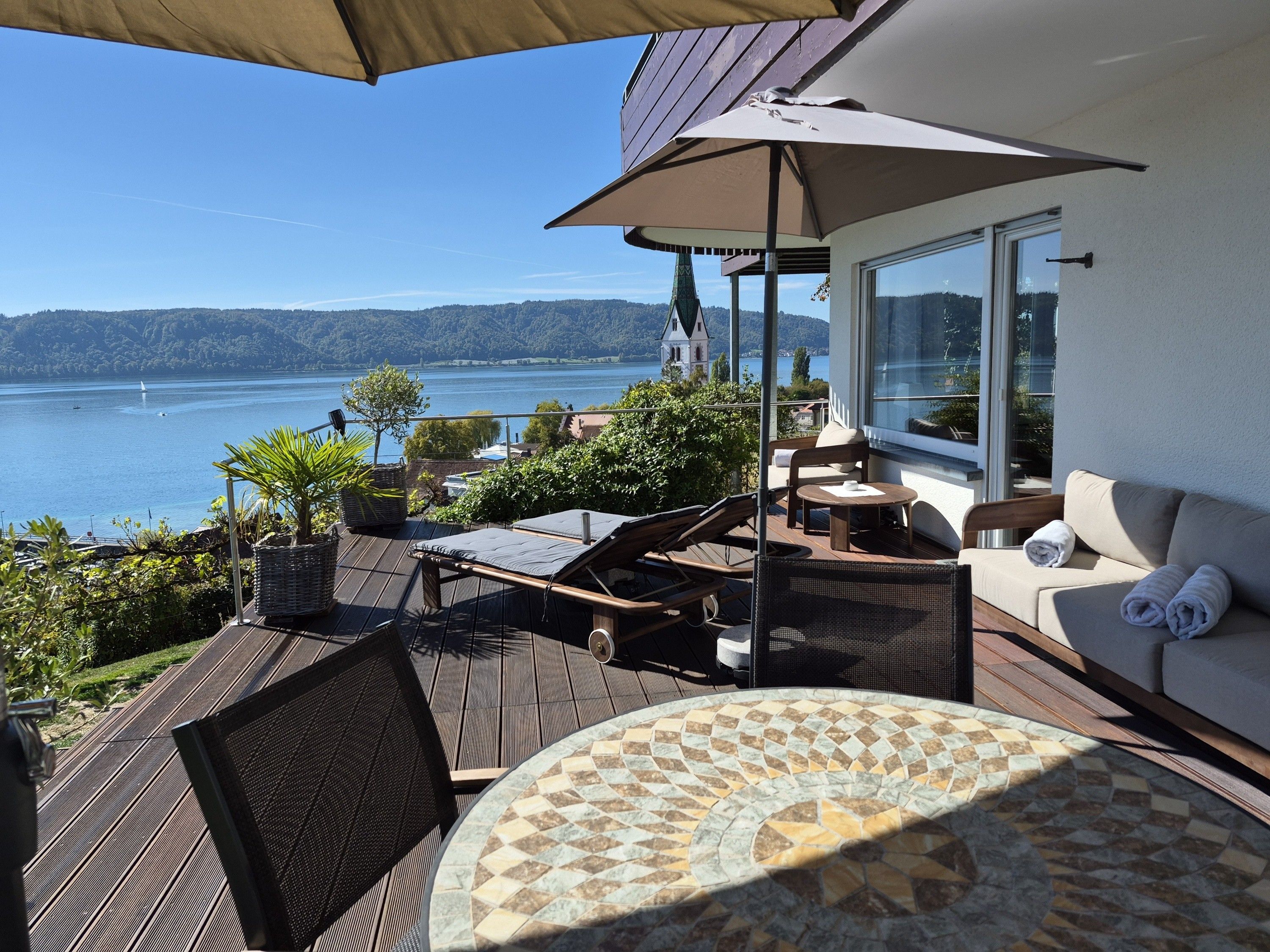 Ein Balkon mit Seeblick, ein Tisch mit kariertem Tischlein und eine Couch mit Handtüchern.