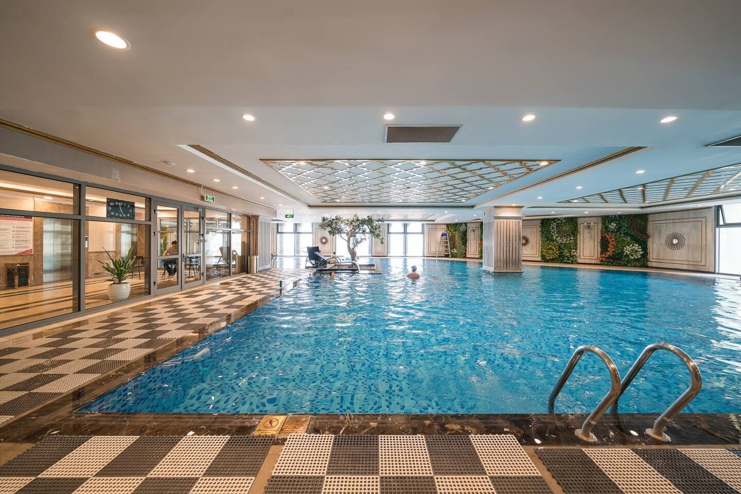 An indoor swimming pool with clear water, surrounded by a checkered floor pattern, and a glass wall on one side.