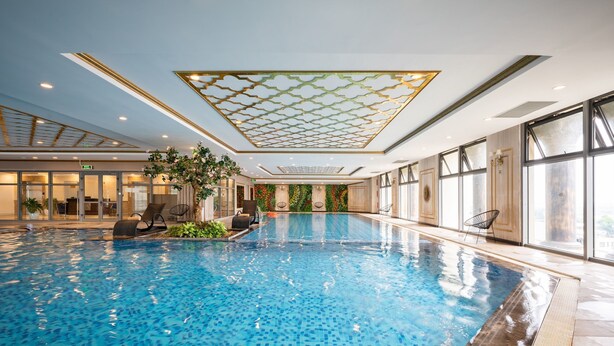 An indoor swimming pool with clear water, a ceiling with a geometric pattern, and a view of the outside through large windows.