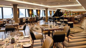 A spacious restaurant with high ceilings, large windows, and a grand piano. Tables are set with fine dining ware.