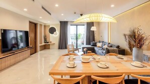 A modern living room with a flat-screen TV, a dining area with a wooden table set for two, and a sofa in the background.