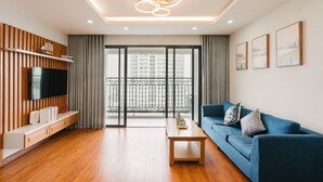 A modern living room with a flat-screen TV, wooden paneling, a blue sofa, a coffee table, and a balcony with a view of high-rise buildings.