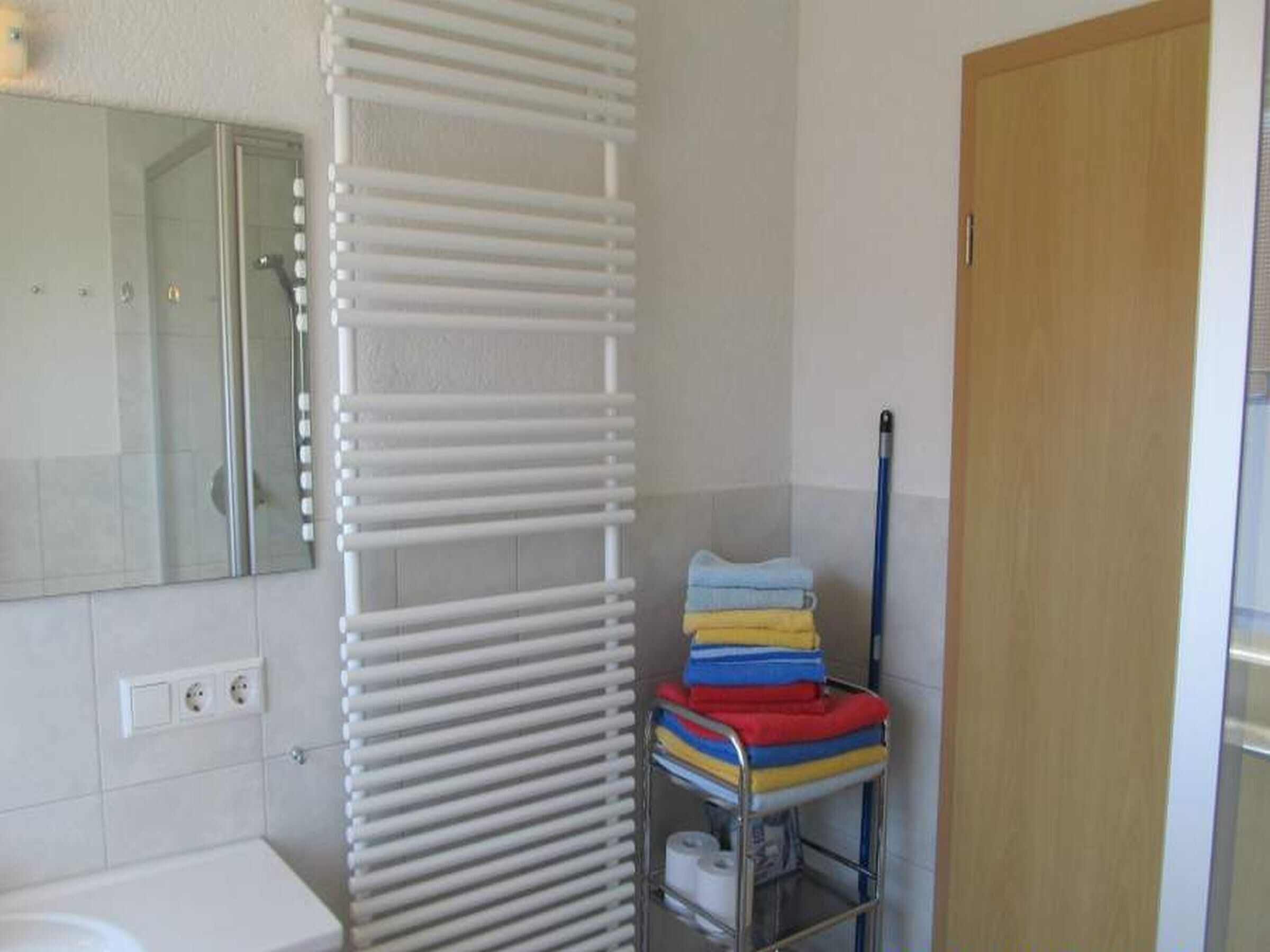 Een badkamer met een witte handdoekhouder, een stapel opgevouwen handdoeken, een metalen plank met schoonmaakspullen en een houten deur.