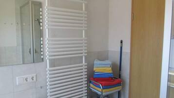 Een badkamer met een witte handdoekhouder, een stapel opgevouwen handdoeken, een metalen plank met schoonmaakspullen en een houten deur.