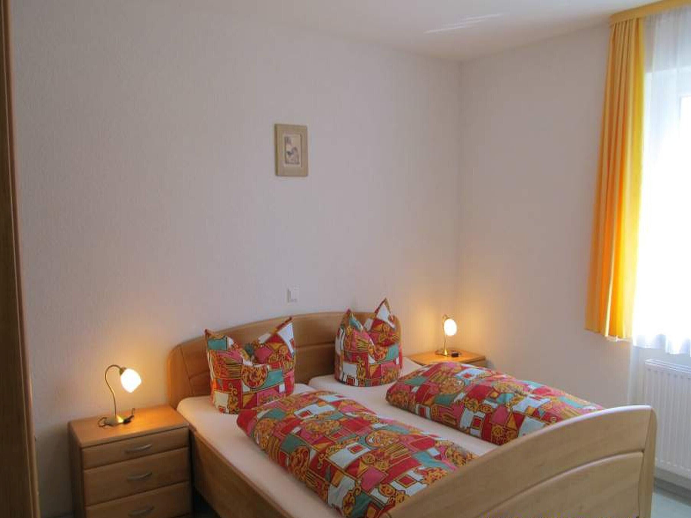 Ein Schlafzimmer mit zwei Betten, einem Nachttisch mit Lampe, einem kleinen Bild an der Wand und einem Fenster mit Vorhängen.