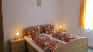 Ein Schlafzimmer mit zwei Betten, einem Nachttisch mit Lampe, einem kleinen Bild an der Wand und einem Fenster mit Vorhängen.