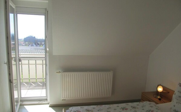 A bedroom with a radiator, a window with a view of a parking lot, and a bedside table with a lamp.