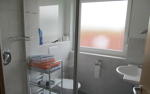 A bathroom with a shower, toilet, and sink. There is a towel rack with folded towels and a bottle of shampoo.