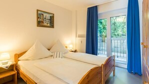 Een tweepersoonsbed met witte lakens, een nachtkastje met een lamp, een schilderij aan de muur en uitzicht op groen buiten het raam.