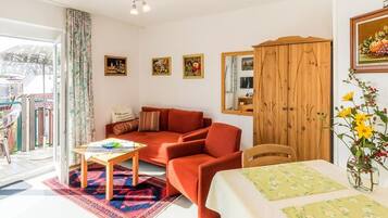 Ein Wohnzimmer mit einer Couch, einem Holzkabinett, einem Esstisch mit floralem Tischläufer und einem Blumenstrauß.