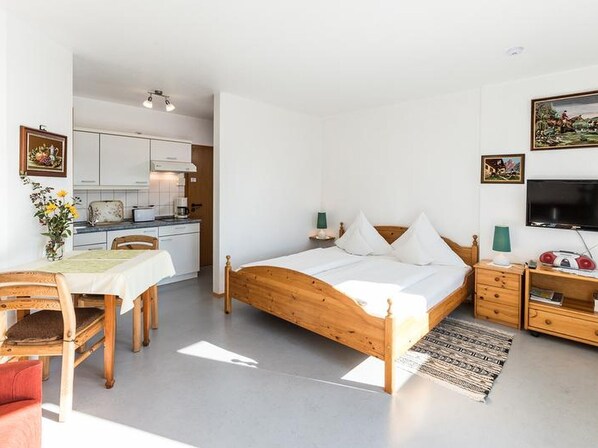Un dormitorio con cama de madera, mesitas de noche, televisor y una zona de cocina con armarios blancos y una mesa de comedor.