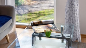 Habitació amb taula de vidre, revista, gerroquet verd i vista a un jardí per la finestra.