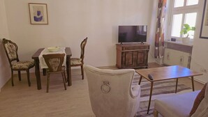 A living room with a dining table, chairs, a sofa, a TV, and a window with curtains. - Holiday Home 'Marienblick' with Private Terrace, Private Garden and Wi-Fi (Beeskow)