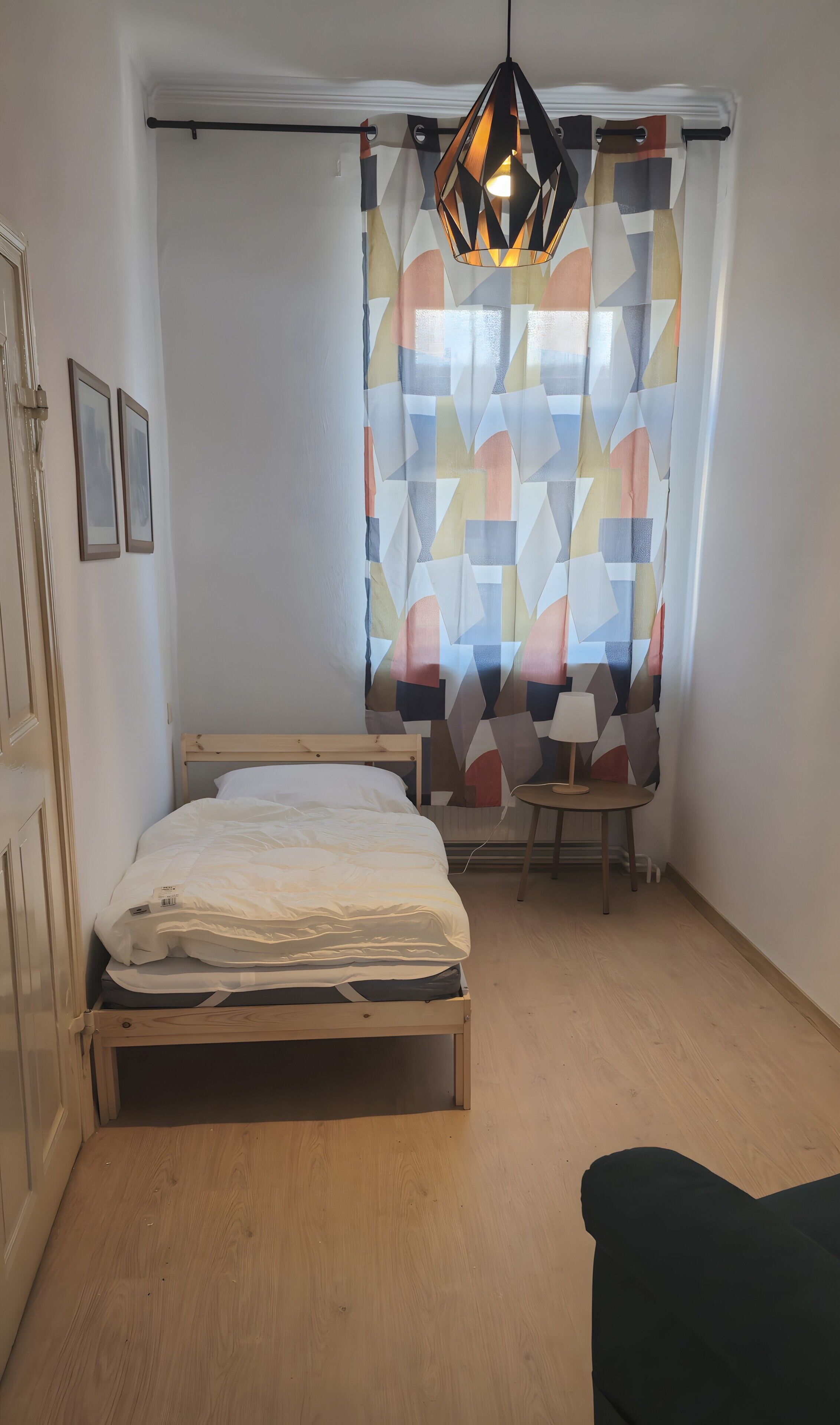 Un dormitorio con cama, mesita de noche, lámpara, lámpara colgante y cortina con estampado geométrico.