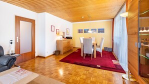 Un salon moderne avec un plafond en bois, un coin repas agrémenté d’un tapis rouge et un meuble vitrine en verre.