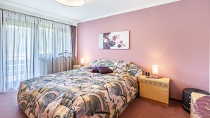 Une chambre à coucher avec un grand lit, une table de chevet avec une lampe, une œuvre d’art accrochée au mur et une fenêtre avec des rideaux.