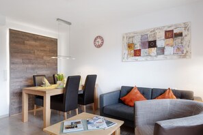 A modern living room with a dining area, a grey sofa with orange cushions, a wooden dining table, and a wall-mounted clock.