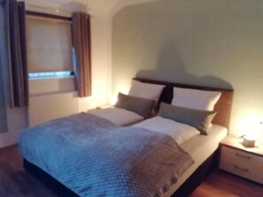 A neatly made bed with pillows and a blanket, a bedside table with a lamp, and a window with curtains.