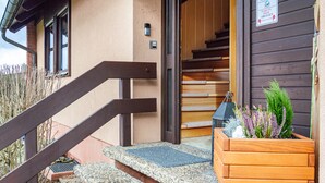 Un escalier en bois menant à l’entrée d’un immeuble, un petit bac à fleurs en bois contenant des plantes et une boîte de dépôt DHL fixée au mur.