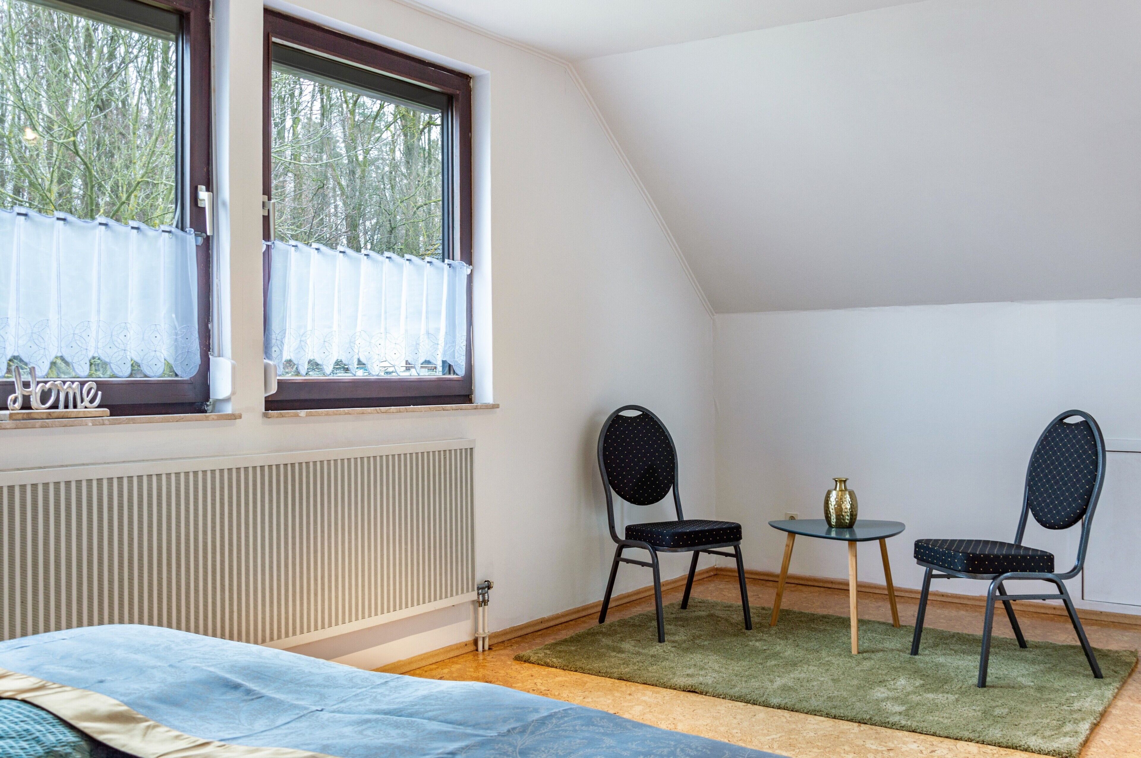 Una habitación con cama, ventana con cortinas, radiador, dos sillas, una mesita y un jarrón.