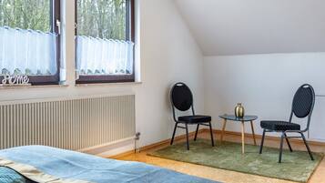 Una habitación con cama, ventana con cortinas, radiador, dos sillas, una mesita y un jarrón.