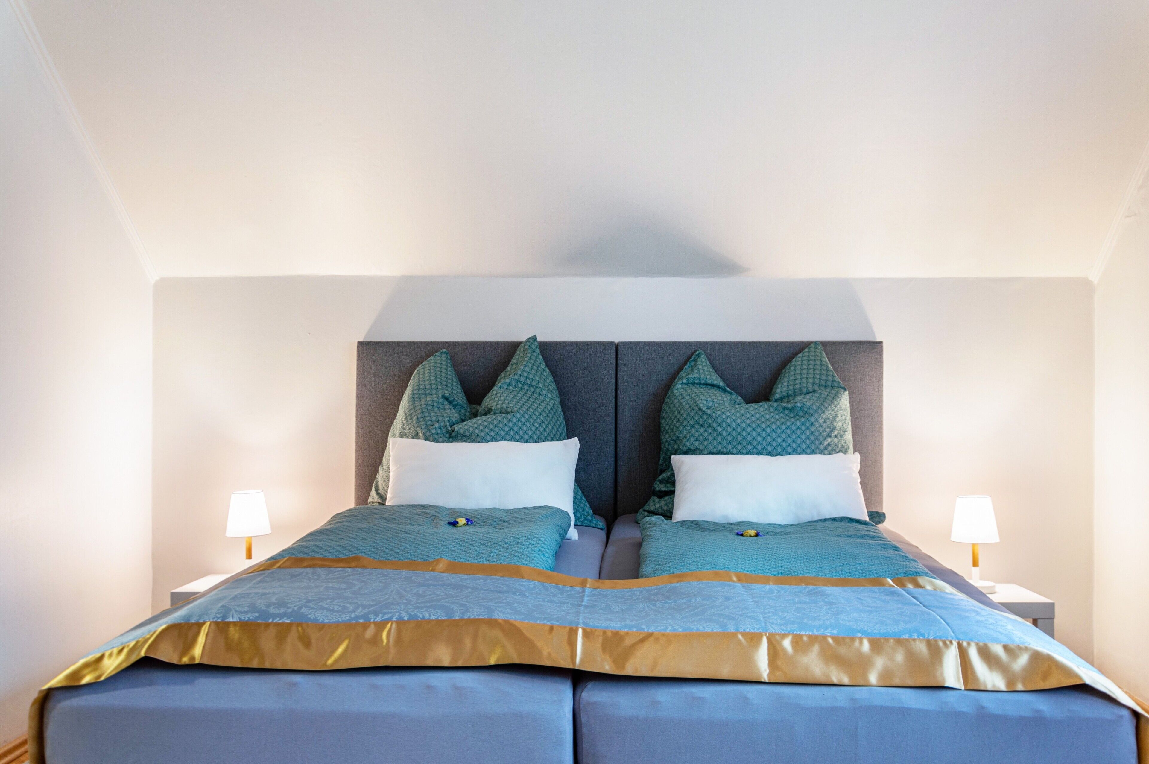 A double bed with a grey headboard, two bedside lamps, and a blue and gold bedspread.