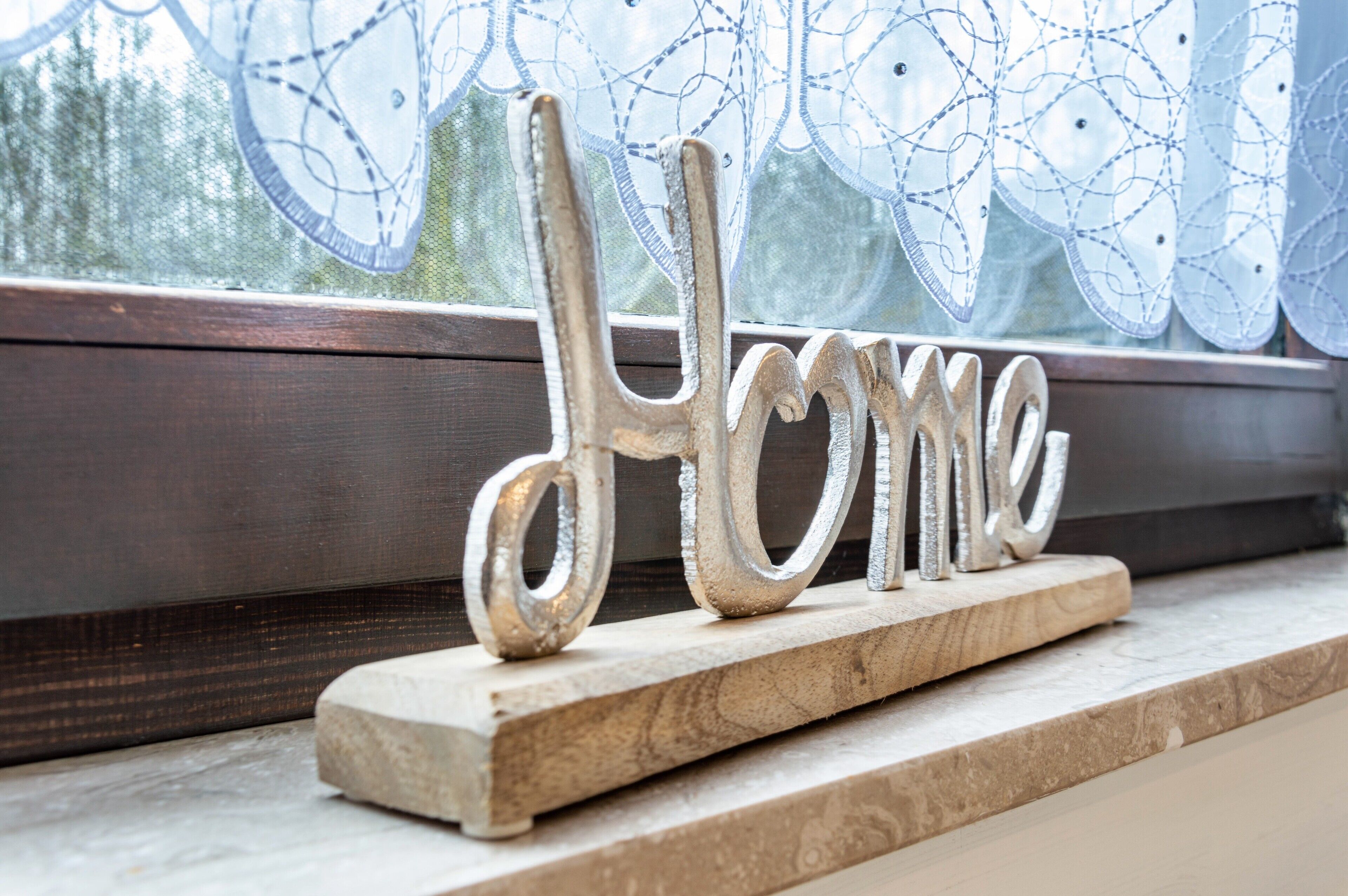 Un cartello decorativo in metallo con la scritta 'Home' su una base di legno vicino a una finestra con tende in pizzo.