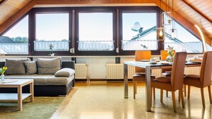 Une pièce mansardée chaleureuse avec un plafond en bois, une table à manger avec des chaises, un canapé et de grandes fenêtres donnant sur une maison.