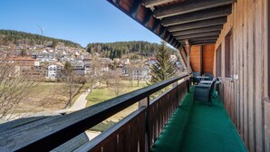 A balcony with a green carpet, wooden railing, and a view of a town with houses and a wind turbine. - Apartment 'Karin' with Lake View and Balcony (Schonach)