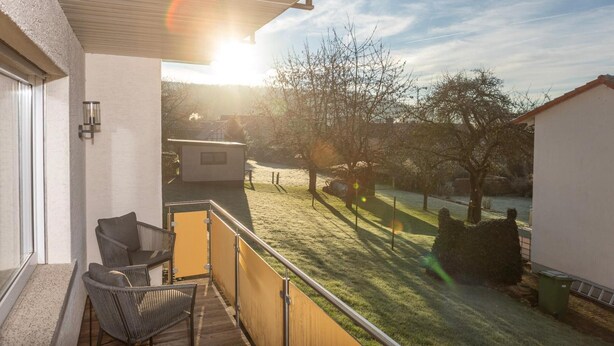 Un balcón con vistas a un jardín césped y árboles, una silla y un edificio pequeño al fondo.