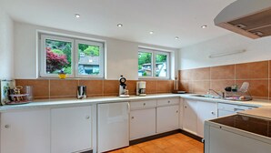 Een keuken met witte kasten, een fornuis, een spoelbak en twee ramen met uitzicht op groen.