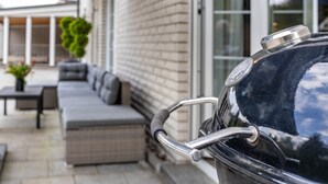 Un barbecue avec un manche en métal, un mur en briques et un espace extérieur aménagé avec des coussins, protégé par un auvent.