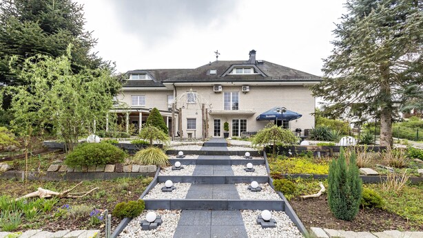 Une grande maison à deux étages, dotée d’un jardin symétrique, d’un chemin central et d’un petit étang.