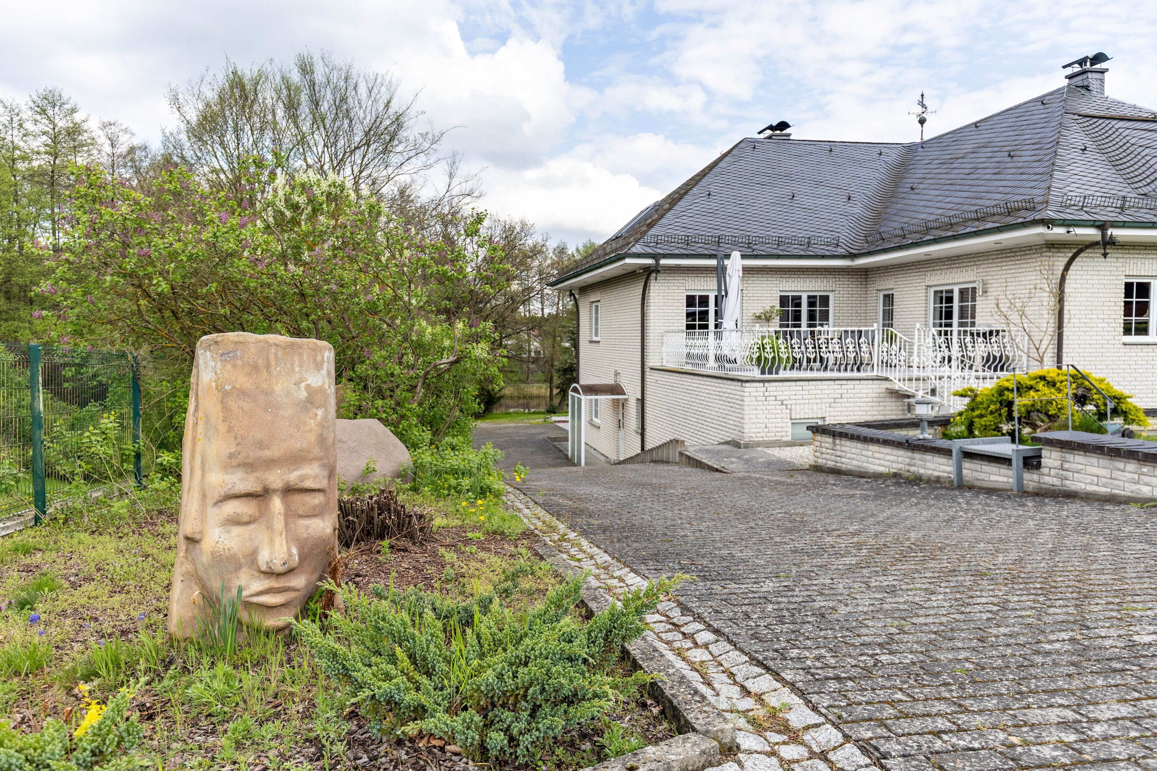 A house with a paved driveway, a white fence, and a garden with a stone sculpture.