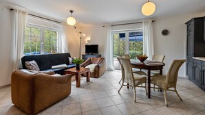 Un salon comprenant un canapé, une table basse, une table à manger avec des chaises, un téléviseur et une vue sur l’extérieur grâce à de grandes fenêtres.