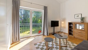 Una habitación con un gran ventanal corredero a un balcón, un televisor de pantalla plana, una mesita redonda y un armario de madera.