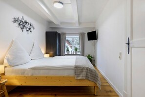 Un dormitorio con cama de madera, paredes blancas, luz de techo, televisor y ventana con cortinas.