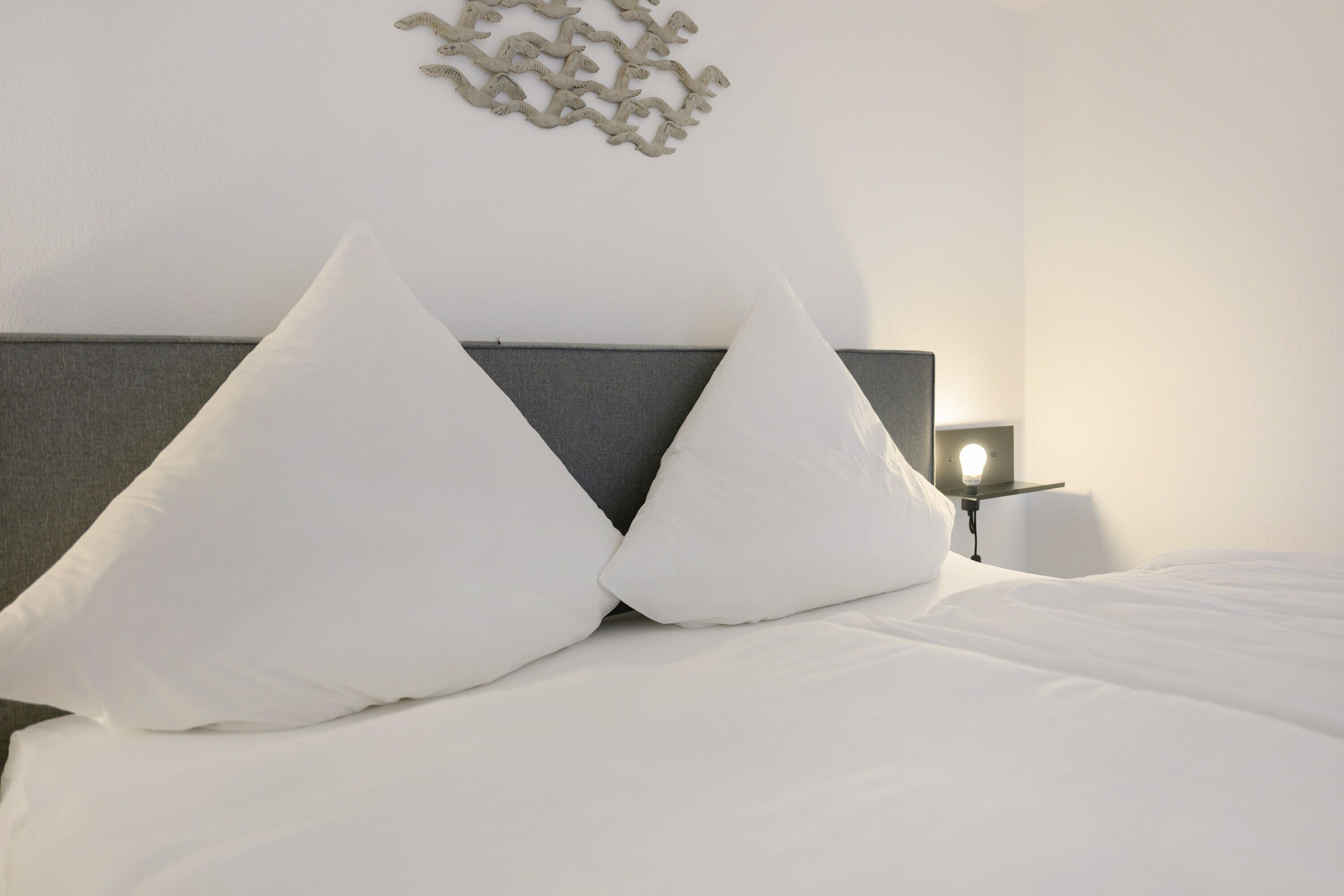 A neatly made bed with white linens and two large pillows, a bedside table with a lamp, and a wall-mounted decorative piece.