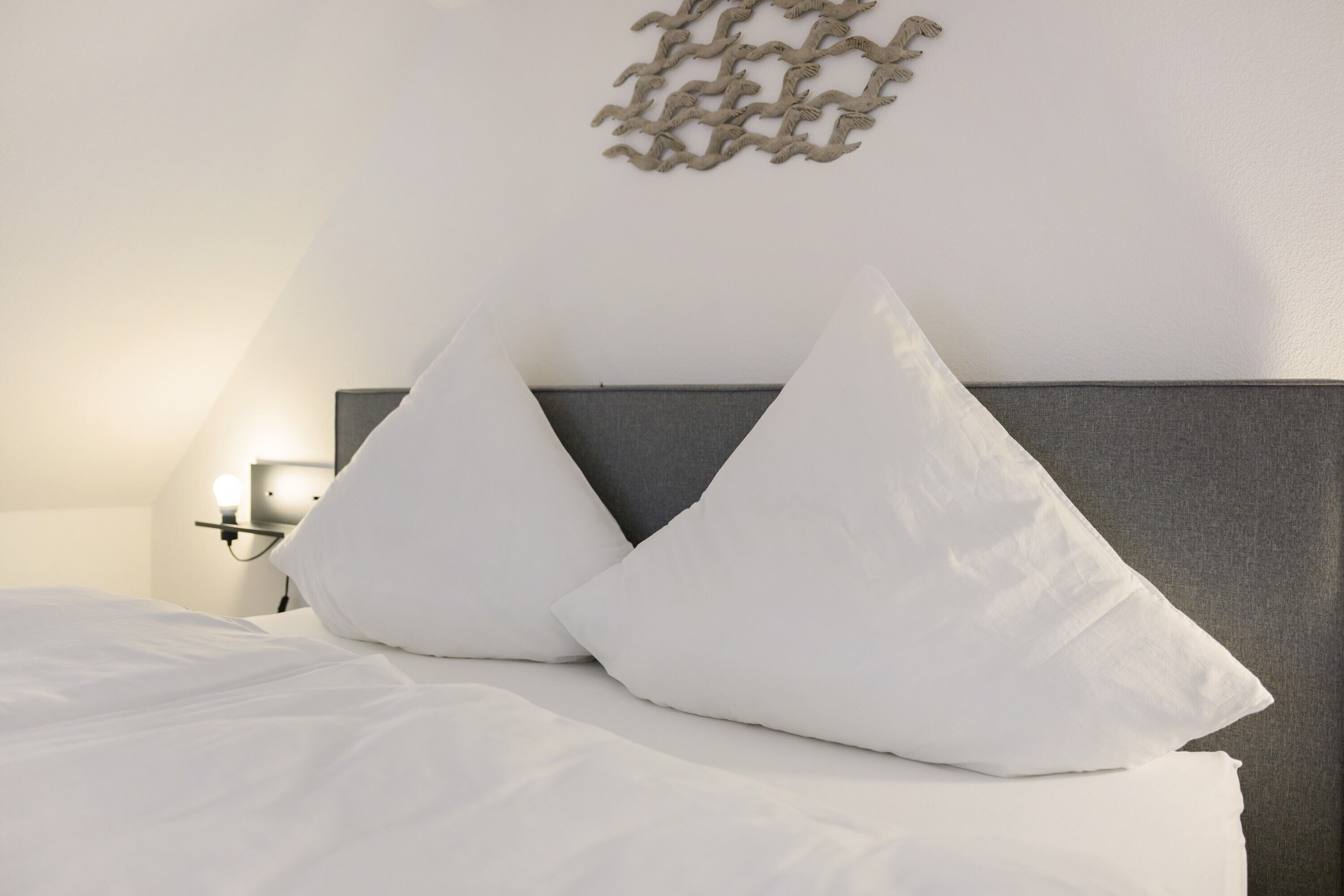 A neatly made bed with white bedding and two white pillows, a gray headboard, and a wall-mounted light fixture.