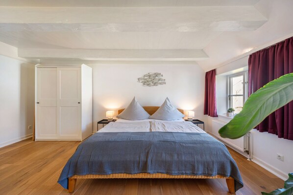 A bedroom with a bed, bedside tables, a window with curtains, a radiator, and a door.