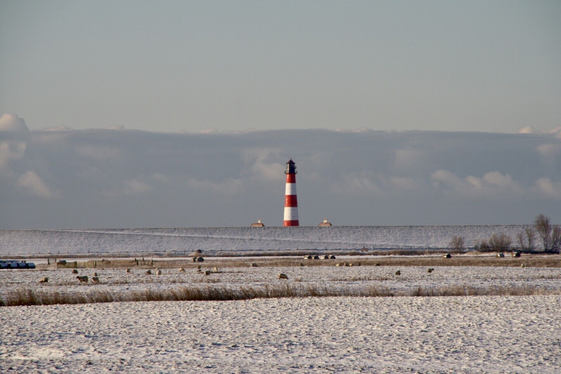 A lighthouse in the distance with a snowy foreground and a flat landscape.