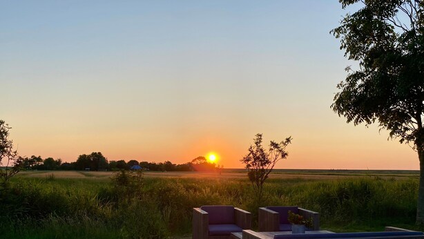 Une terrasse en bois, agrémentée de sièges et de plantes en pot, qui offre une vue sur un champ et sur le soleil couchant à l’horizon.