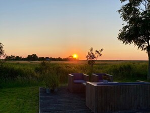 En träveranda med sittplatser och krukväxter, med utsikt över ett fält och en solnedgång i fjärran.